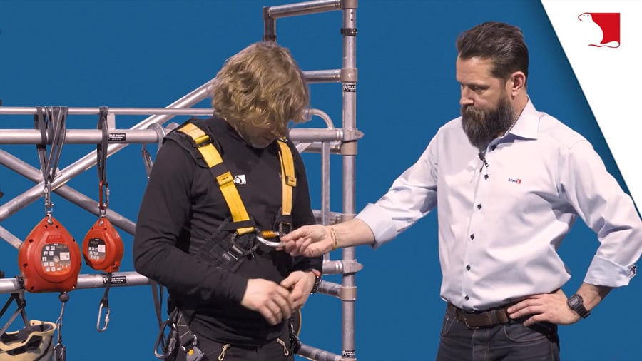 Två män vid en arbetsstation med säkerhetsutrustning och sele, en i vit skjorta och en i svart tröja med gul säkerhetssele. Bakgrunden visar metallställningar och räddningsutrustning.