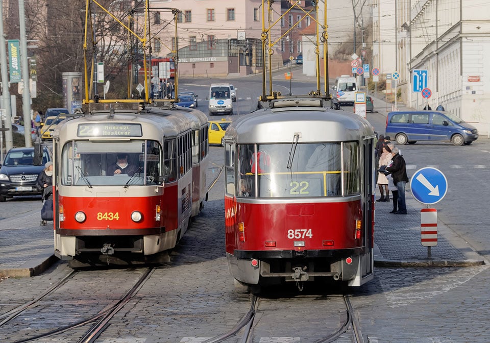 Två röda och grå spårvagnar på en stadsgata med korsande spår och omgivande byggnader i bakgrunden.