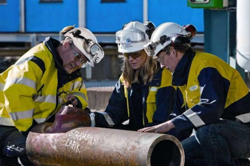 Tre personer från Kiwa i skyddsutrustning inspekterar ett stort metallrör utomhus, med en blå byggnad i bakgrunden.