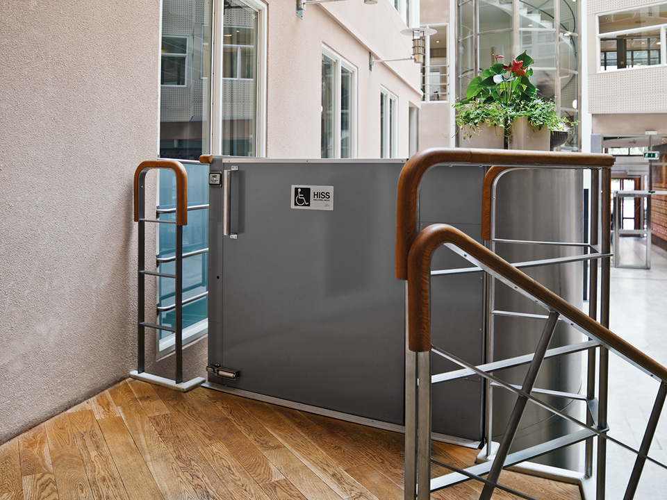 Plattformshiss vid en inomhustrappa med ledstänger i trä och metall i en ljus byggnadsmiljö.