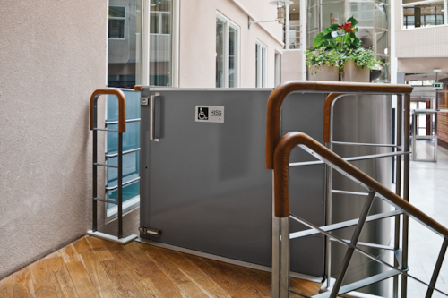 Plattformshiss vid en inomhustrappa med ledstänger i trä och metall i en ljus byggnadsmiljö.