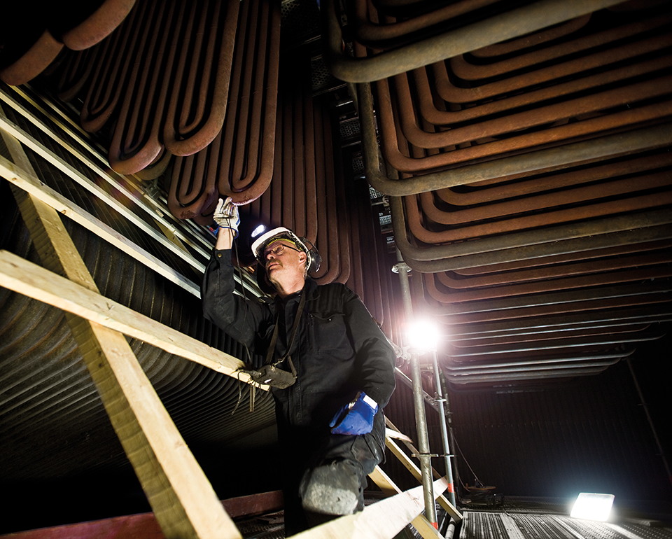 Inspektör i skyddsutrustning kontrollerar metallrör i en industriell anläggning, med byggnadsställningar och belysning i närheten.