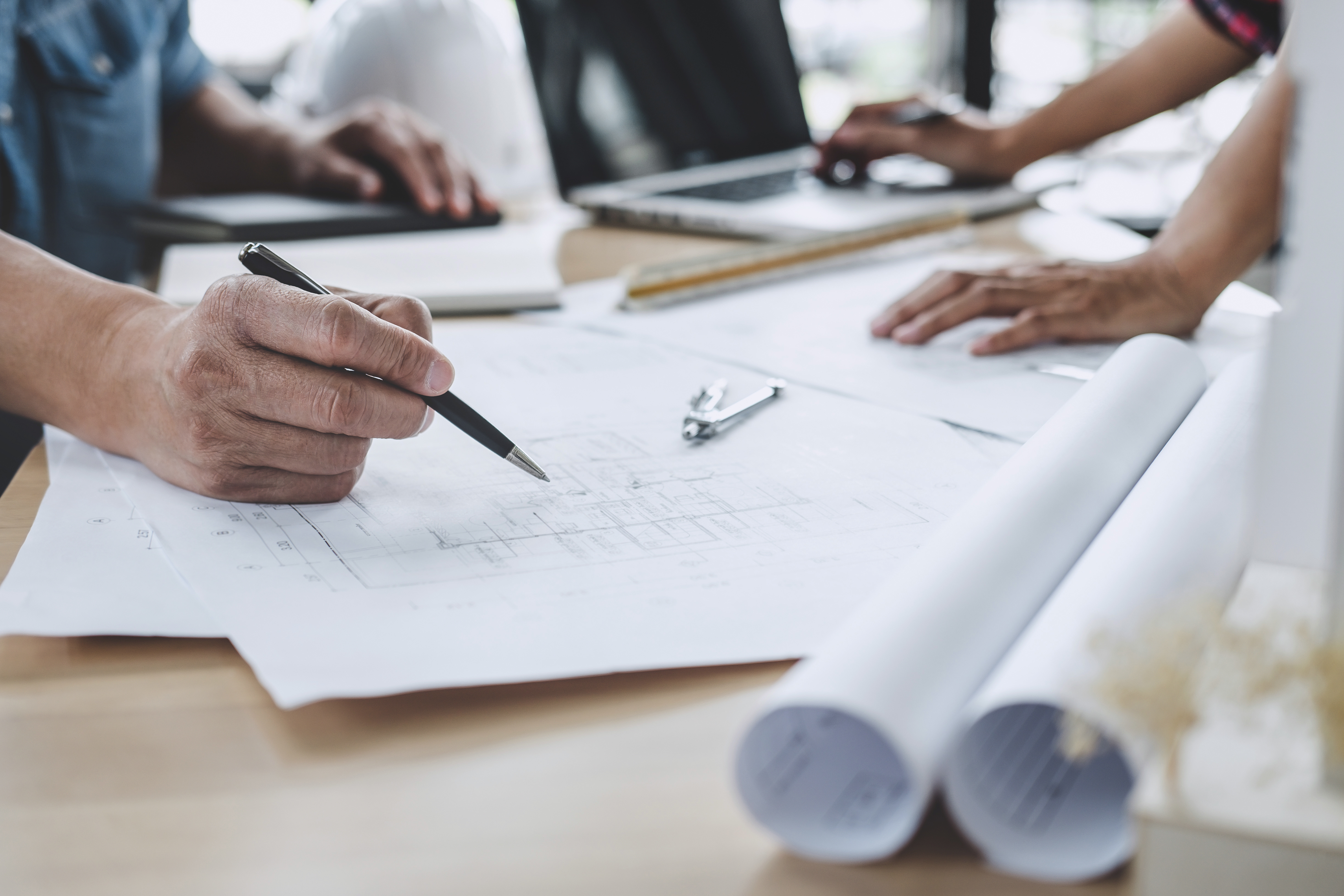 Architect reviewing blueprints at a desk with rolled plans and a clipboard, while the colleague uses a laptop in the background