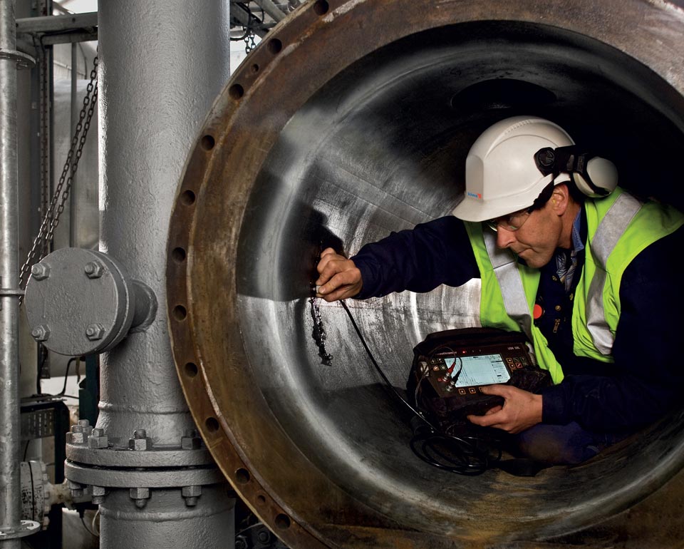 A Kiwa NDT employee wearing a safety vest and helmet operates equipment to inspect the interior of a large industrial pipe.