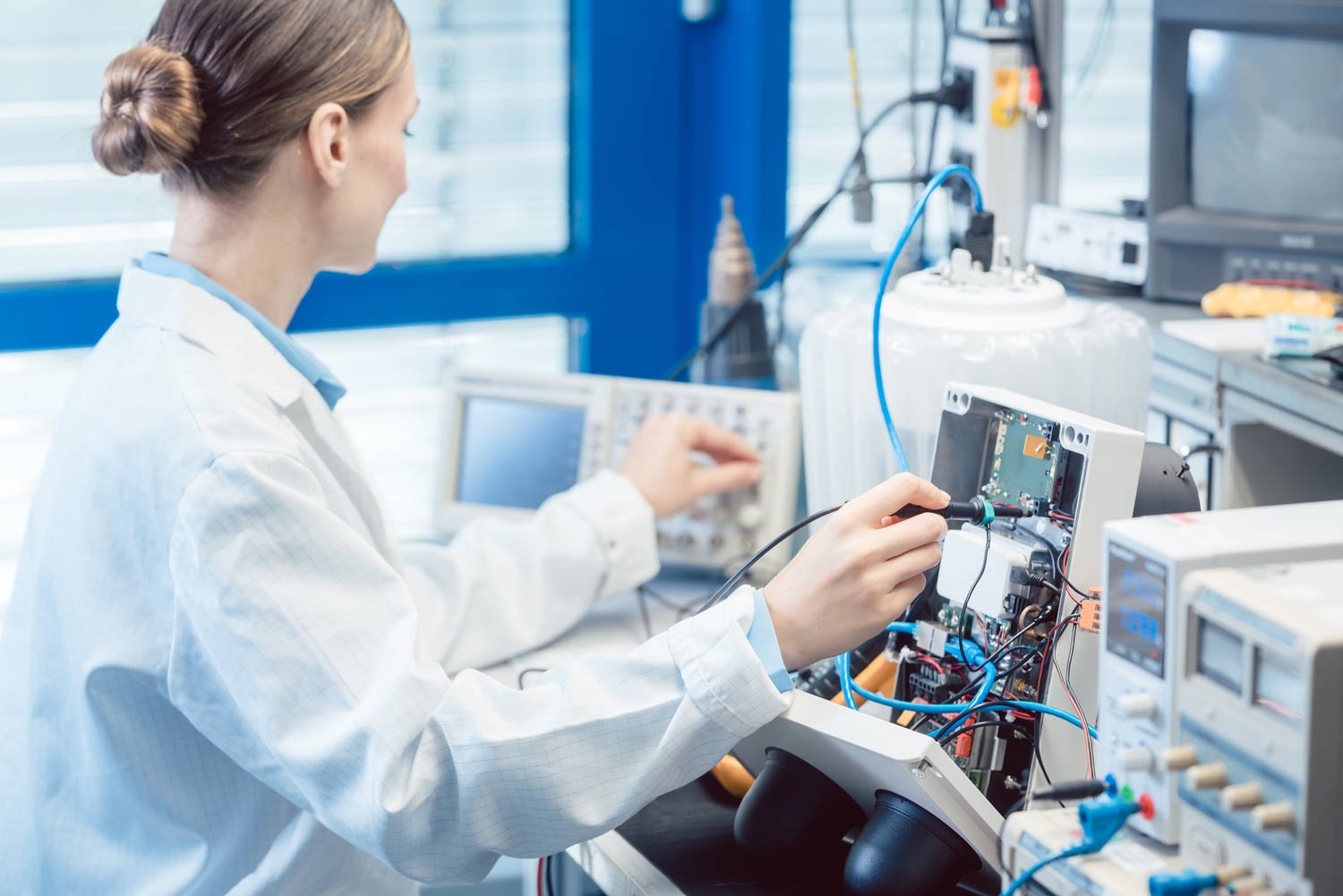 A female Kiwa lab employee wearing a lab coat conducts electronics testing in a laboratory, using advanced oscilloscopes and other technical equipment for research and development
