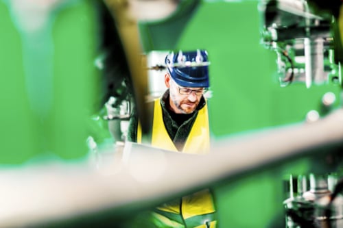 En person i gul säkerhetsväst och blå skyddshjälm omgiven av industriell maskinutrustning, med fokus på mekaniska delar.