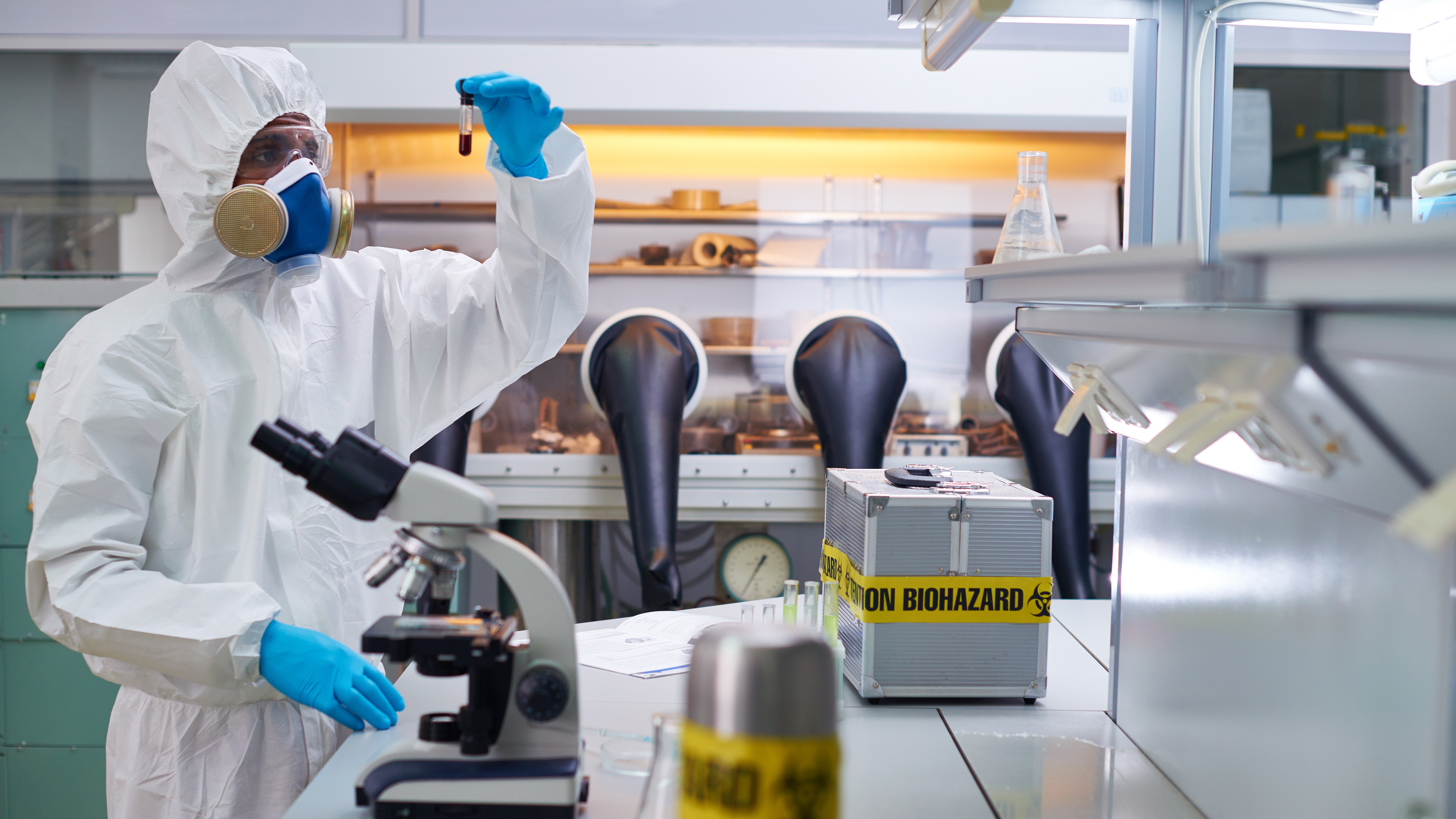 Scientist in a hazmat suit and respirator holding a test tube in a laboratory with a microscope and biohazard container