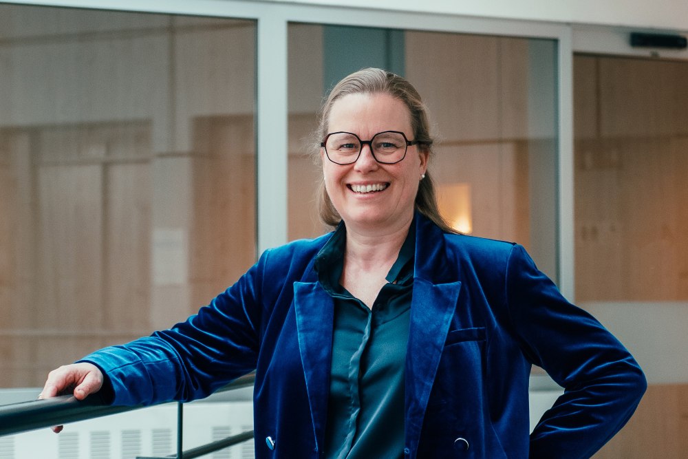 Smiling person wearing glasses and a blue velvet blazer standing indoors near a glass railing