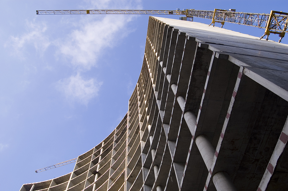 Gebouw in aanbouw met betonnen structuren en verticale lijnen, geflankeerd door bouwkranen tegen een blauwe lucht.