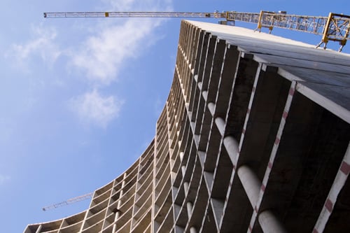 Gebouw in aanbouw met betonnen structuren en verticale lijnen, geflankeerd door bouwkranen tegen een blauwe lucht.