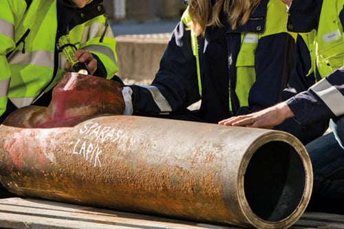 Ingenjörer i varselkläder inspekterar ett stort rostigt metallrör som ligger på en träpall.