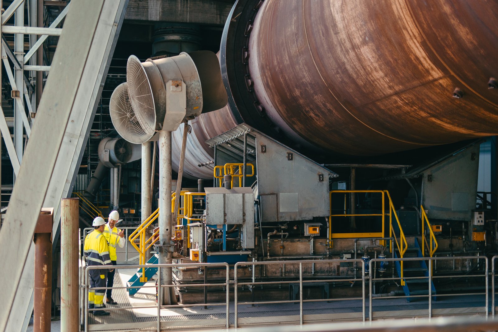 Kiwa auditors in safety gear inspect machinery at a manufacturing plant, focusing on a large, rusted cylindrical structure and ventilation system