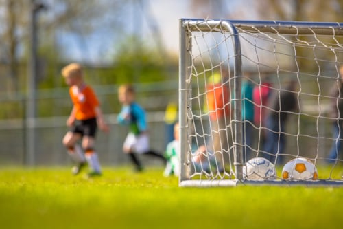 A soccer goal with two balls inside on a grassy field, with children playing soccer in the background.