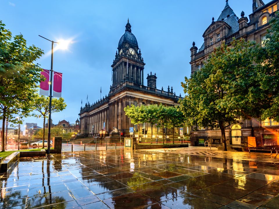 Historisch gebouw met koepel en torens, omringd door bomen, straatverlichting en een natte geplaveide straat tijdens de schemering.
