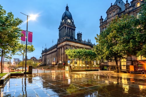 Historisch gebouw met koepel en torens, omringd door bomen, straatverlichting en een natte geplaveide straat tijdens de schemering.