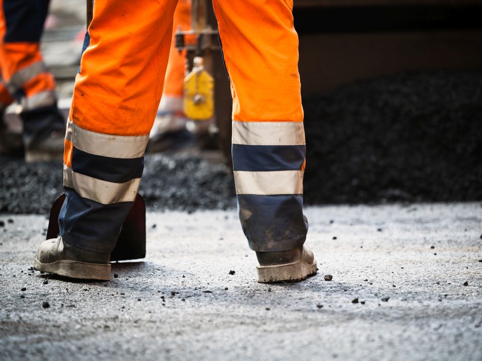 Persoon in oranje werkbroek met reflecterende strepen bij een wegwerkzaamheden, omringd door asfalt en gereedschap.