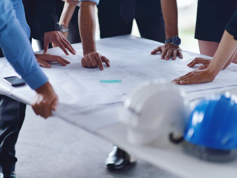 Mensen bekijken en wijzen naar bouwplannen op een tafel, met bouwhelmen en andere accessoires op de voorgrond.