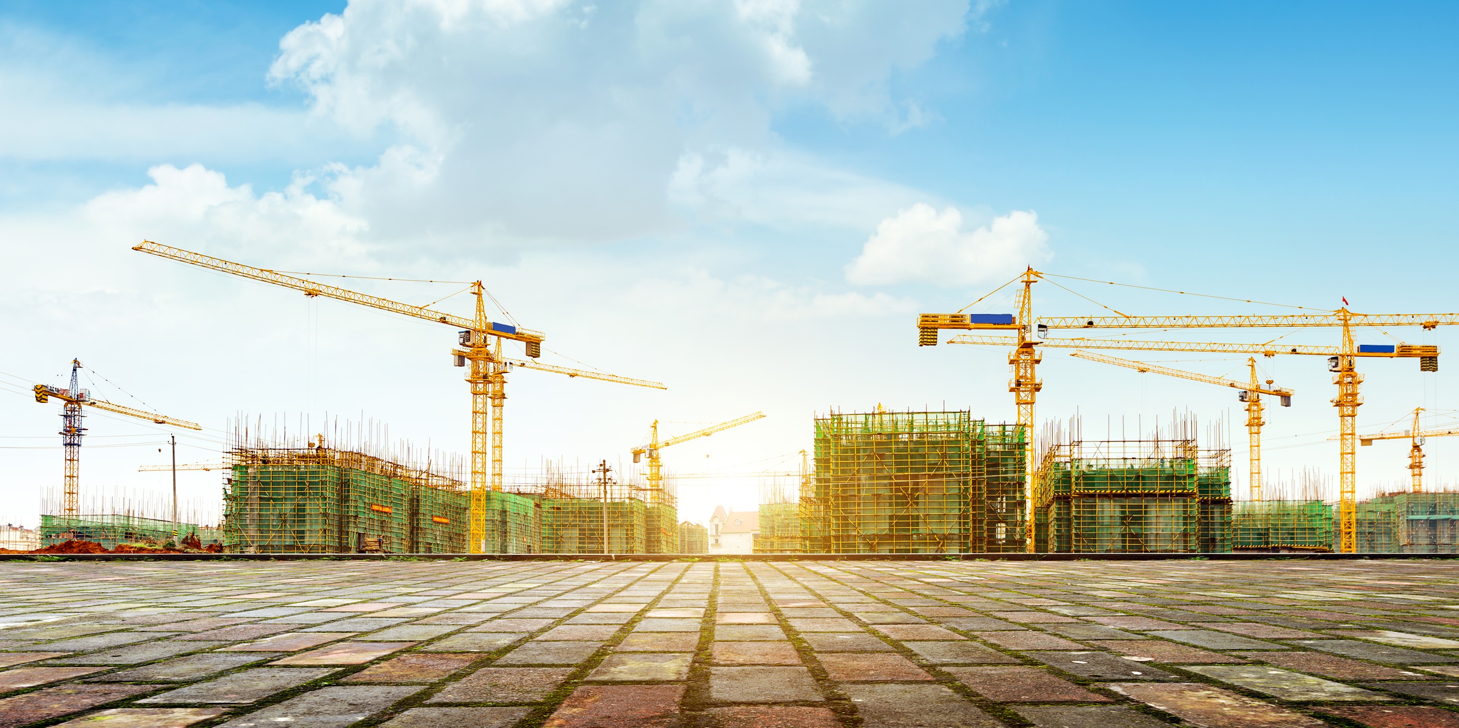 Bouwplaats met meerdere torenkranen en gebouwen in aanbouw, onder een blauwe lucht met witte wolken.