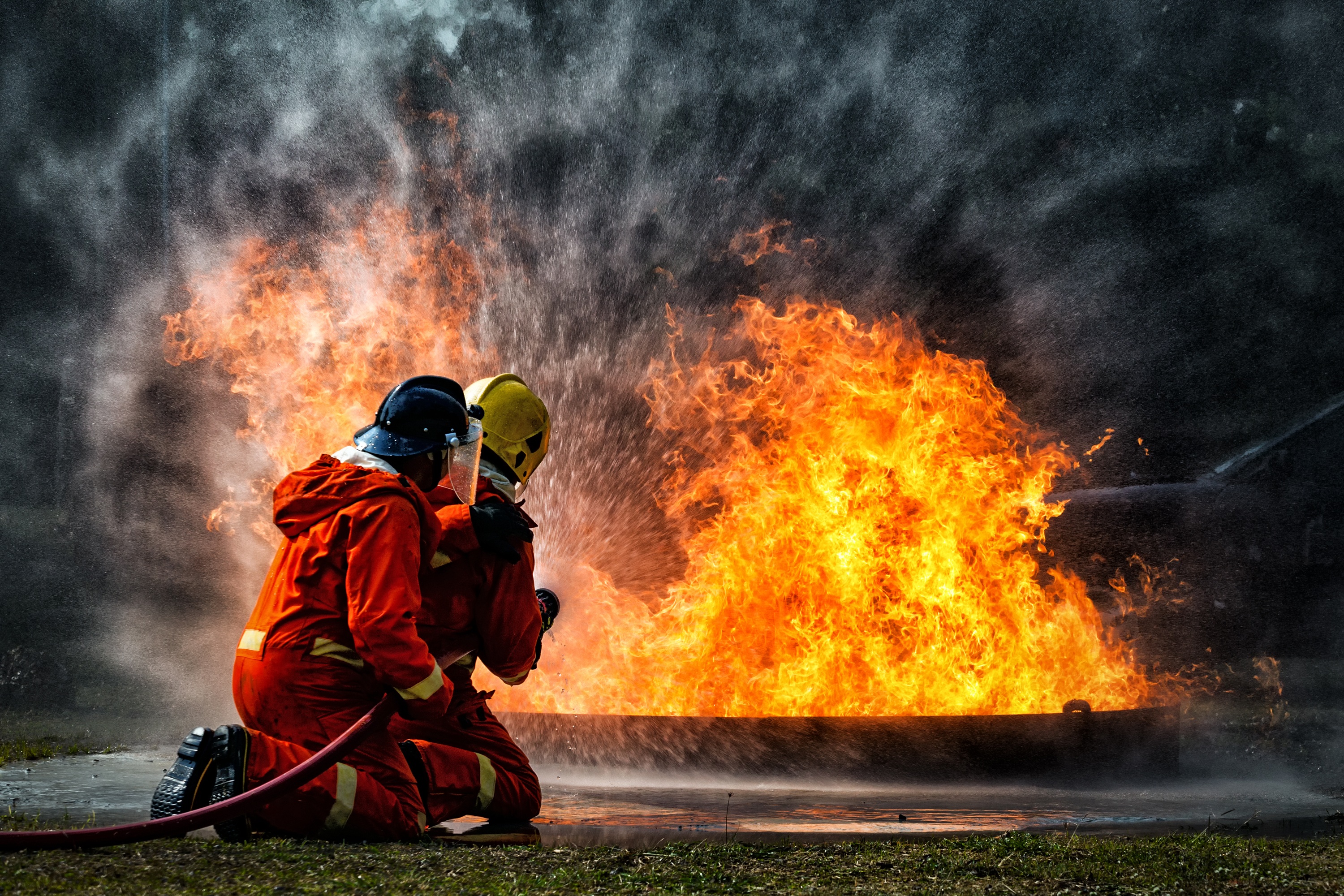 Firefighters in protective gear and helmets kneeling in front of a large blaze 