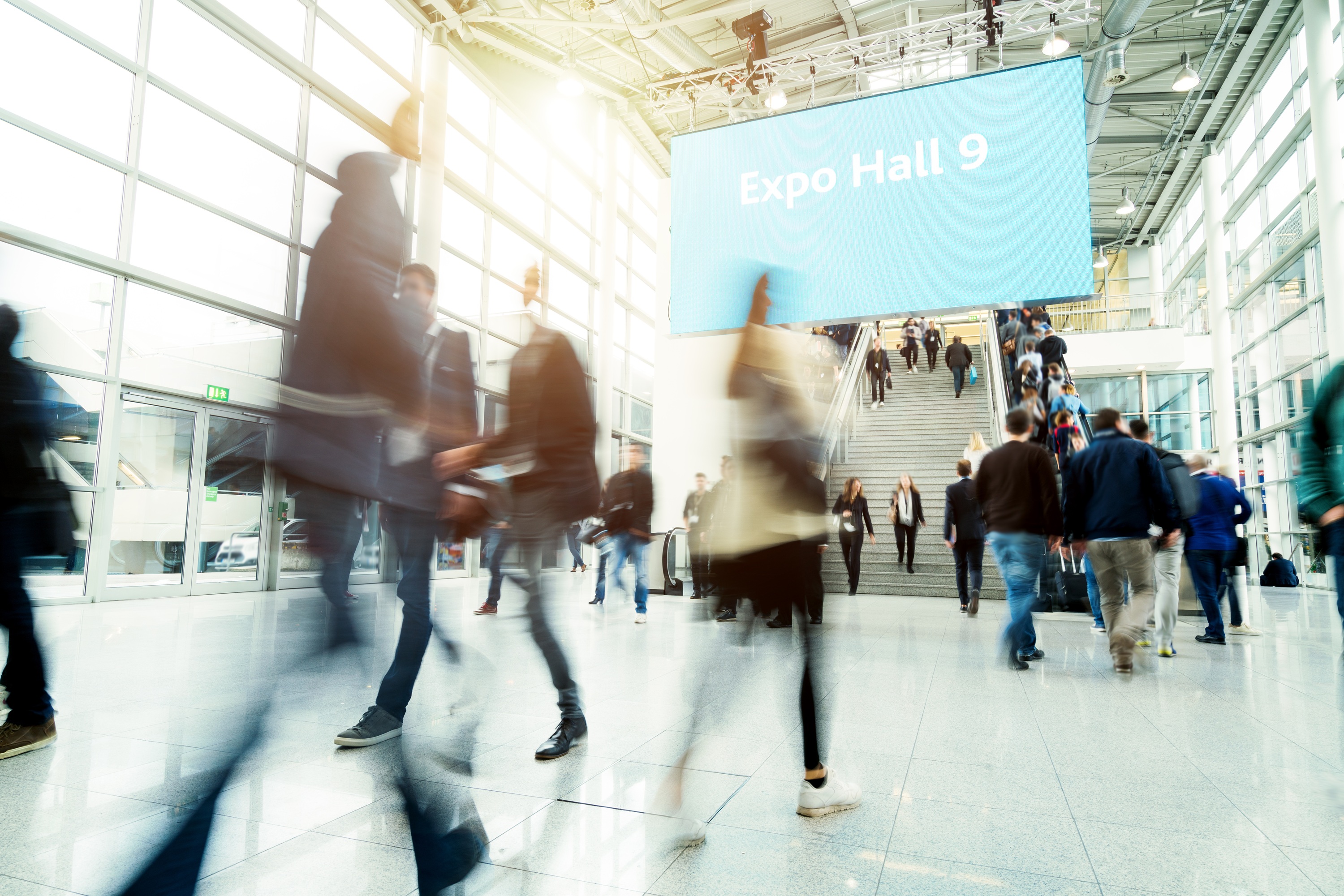 Personen lopen door een lichte, moderne hal met glazen wanden en een groot bord dat "Expo Hall 9" aangeeft bij een trap.