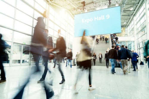 Personen lopen door een lichte, moderne hal met glazen wanden en een groot bord dat "Expo Hall 9" aangeeft bij een trap.