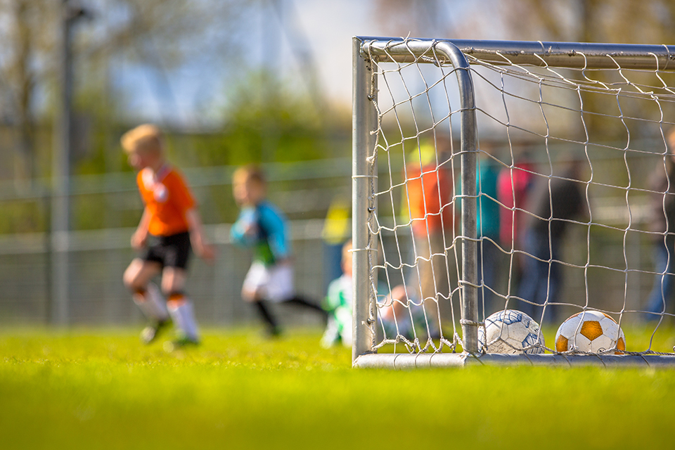 Voetbaldoel met ballen erin op een grasveld, terwijl kinderen op de achtergrond een wedstrijd spelen.