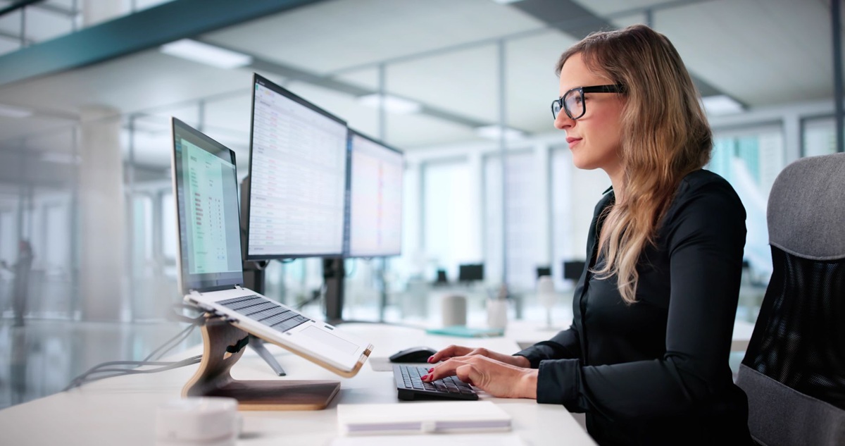 A woman working at a desk with multiple computer monitors in a modern office setting.