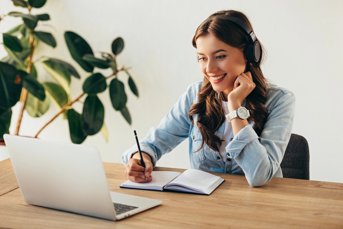 Vrouw zit aan een houten tafel met een laptop en schrijft in een notitieboekje