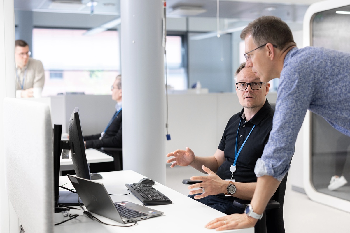 Two men talking in a modern office with desktop computers and laptops on the desk. One man is seated while another leans over to speak with him.