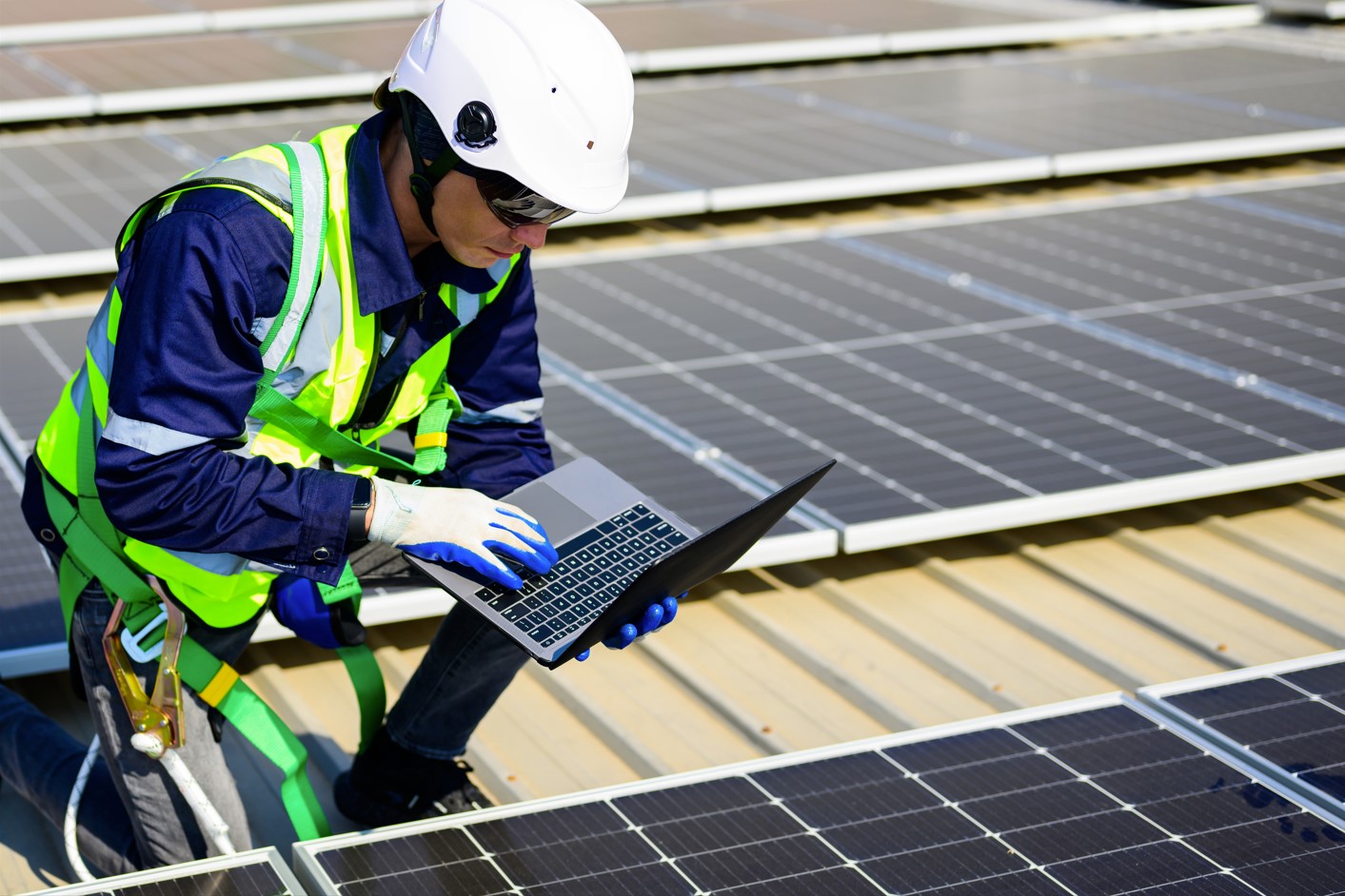 Trabajador con casco y chaleco reflectante inspeccionando paneles solares sobre un tejado mientras utiliza un ordenador portátil.