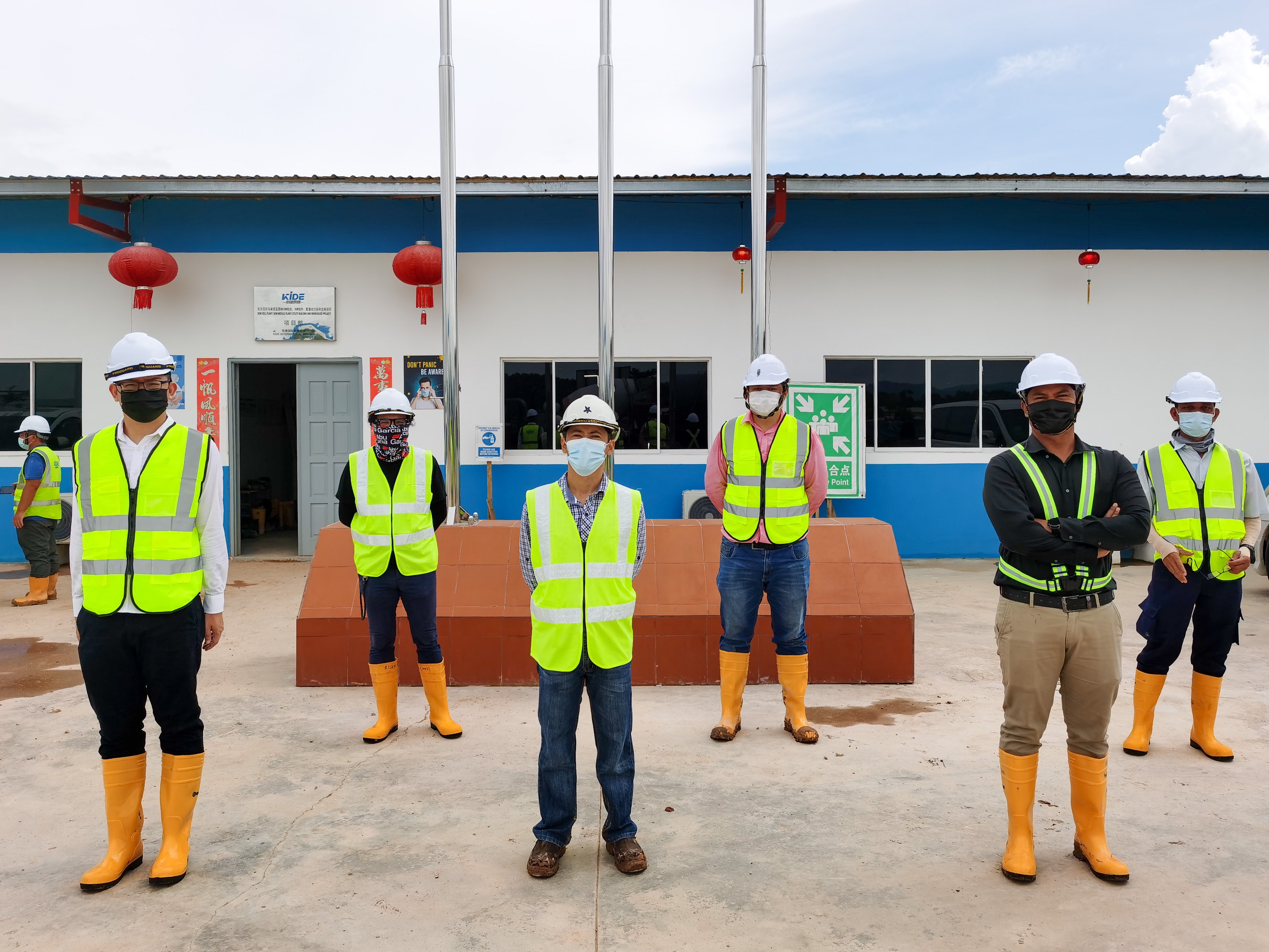 Trabajadores de la construcción con cascos y chalecos reflectantes amarillos, de pie frente a un edificio de obra con botas de seguridad naranjas.