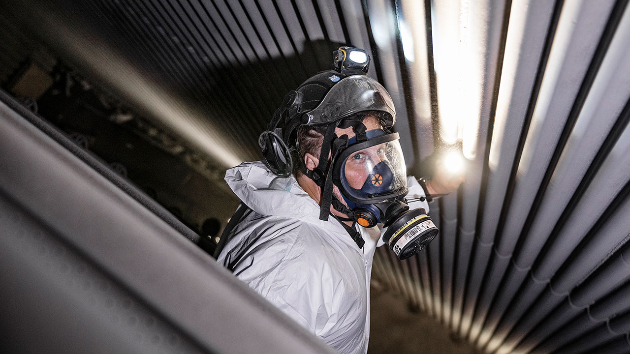 En person i skyddsutrustning, inklusive hjälm och andningsmask, inspekterar metallrör i en industrimiljö med en ficklampa.