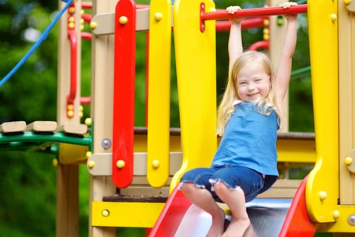 En liten flicka har roligt på en lekplats, hon håller båda händerna i en stång ovanför hennes huvud, hänger lite i den, och står barfota på rutschkanan som hon verkar vilja åka i.