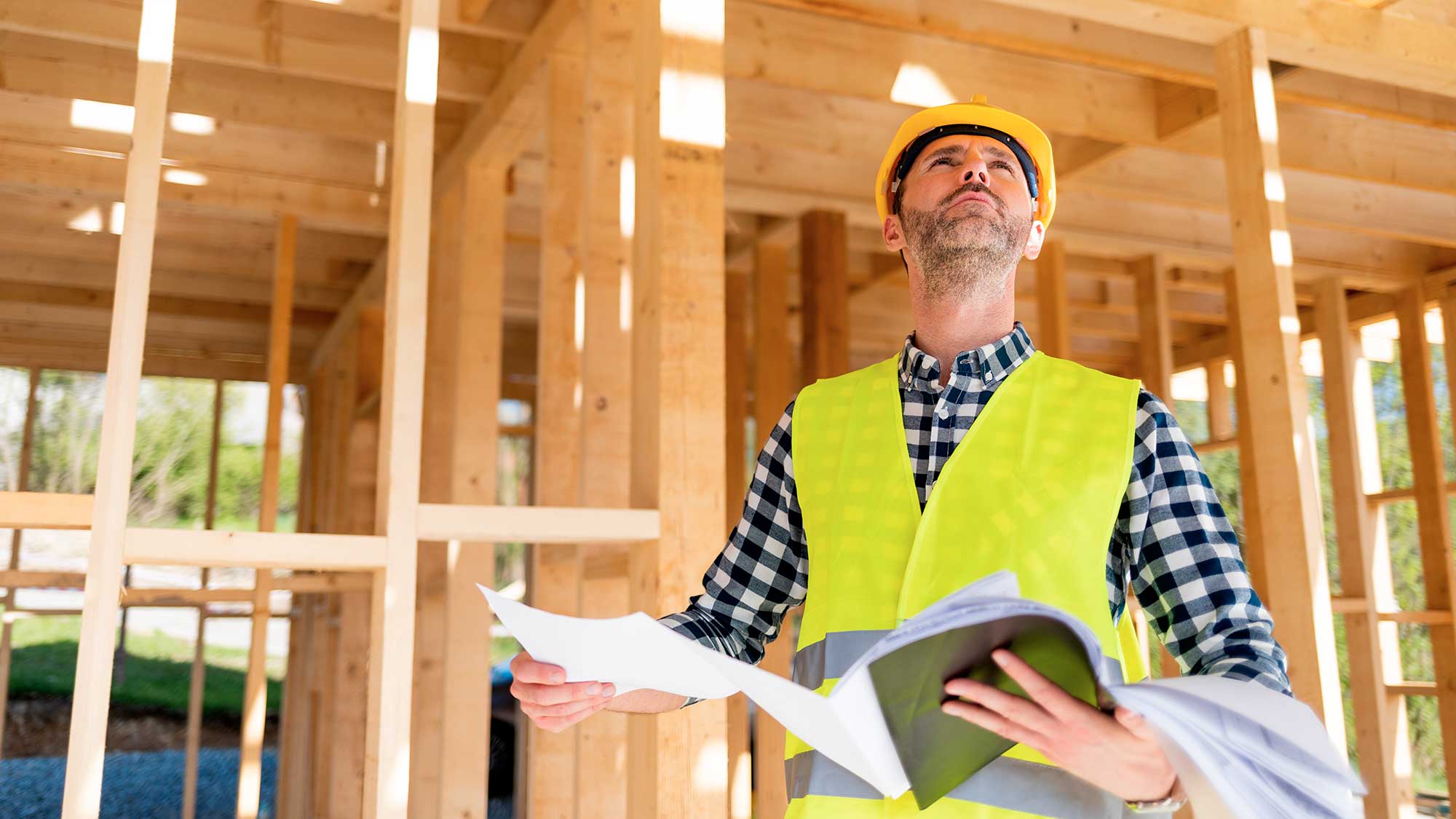 Miljöinventerare i reflexväst och skyddshjälm håller planer på en byggarbetsplats med träkonstruktioner.