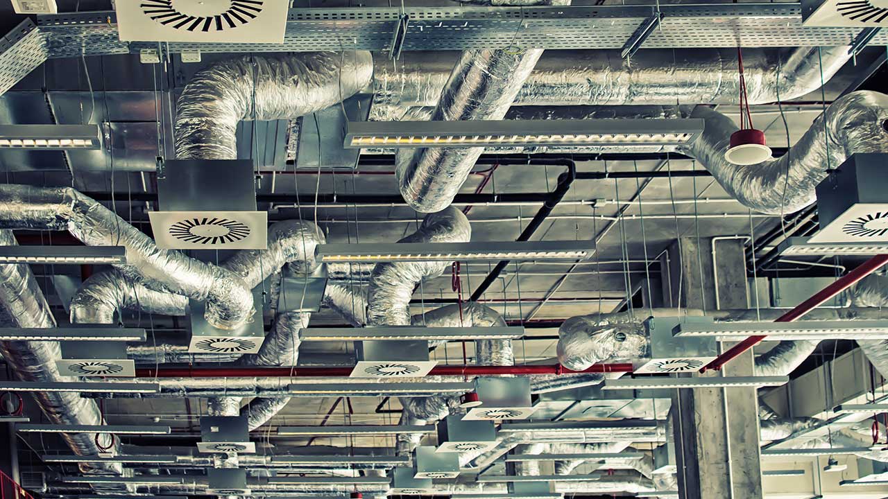 Ventilationssystem uppe i taket med flera rör och fläktar i en industrilokal. 