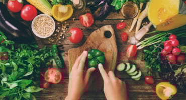 Händer som skär en grön paprika på en träskärbräda, omgiven av färska grönsaker och ingredienser på ett rustikt bord.