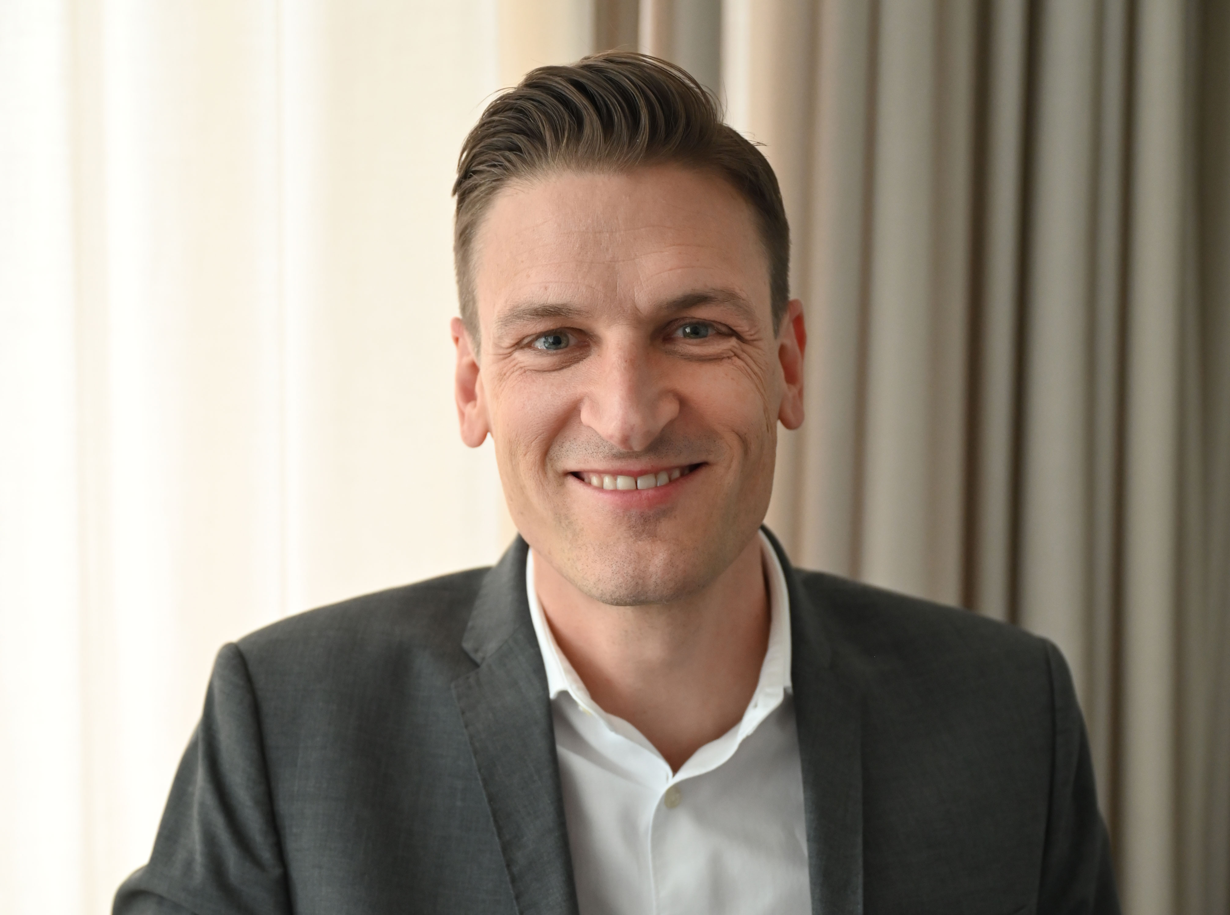 Man i kostym och vit skjorta sitter framför ett neutralt beige draperi.