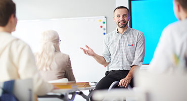 En man i rutig skjorta talar inför en grupp människor i ett klassrum. En whiteboard och en stor blå skärm syns i bakgrunden.