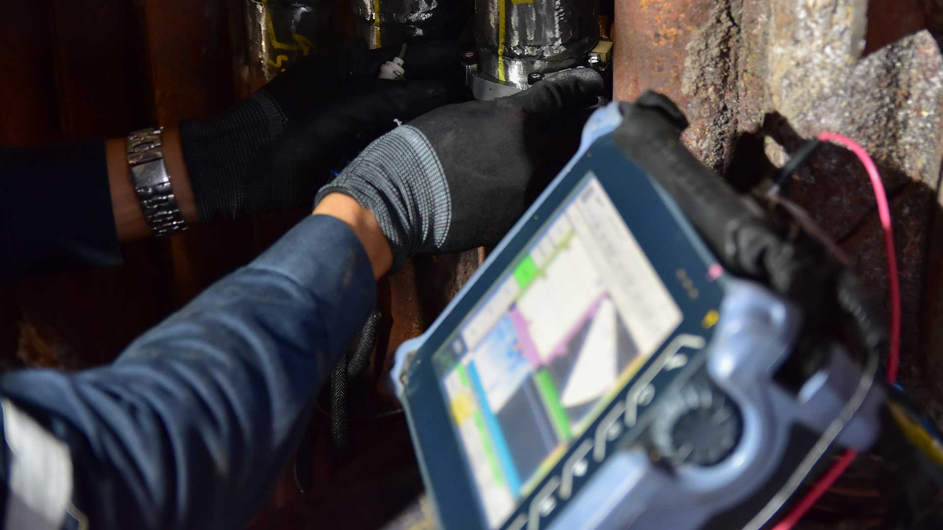 En person använder en handhållen ultraljudsmätare för att inspektera rostiga metallrör.