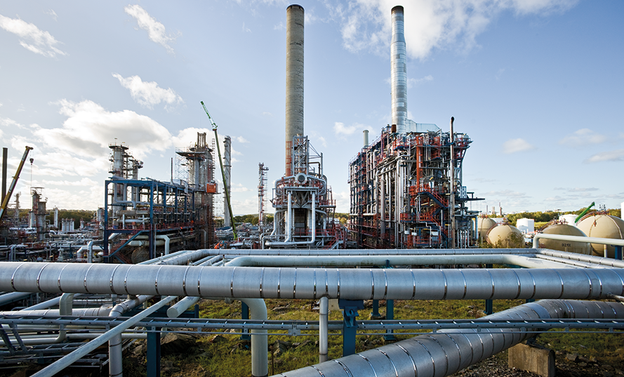 Industriell anläggning med rörledningar, skorstenar och byggnader under en blå himmel.
