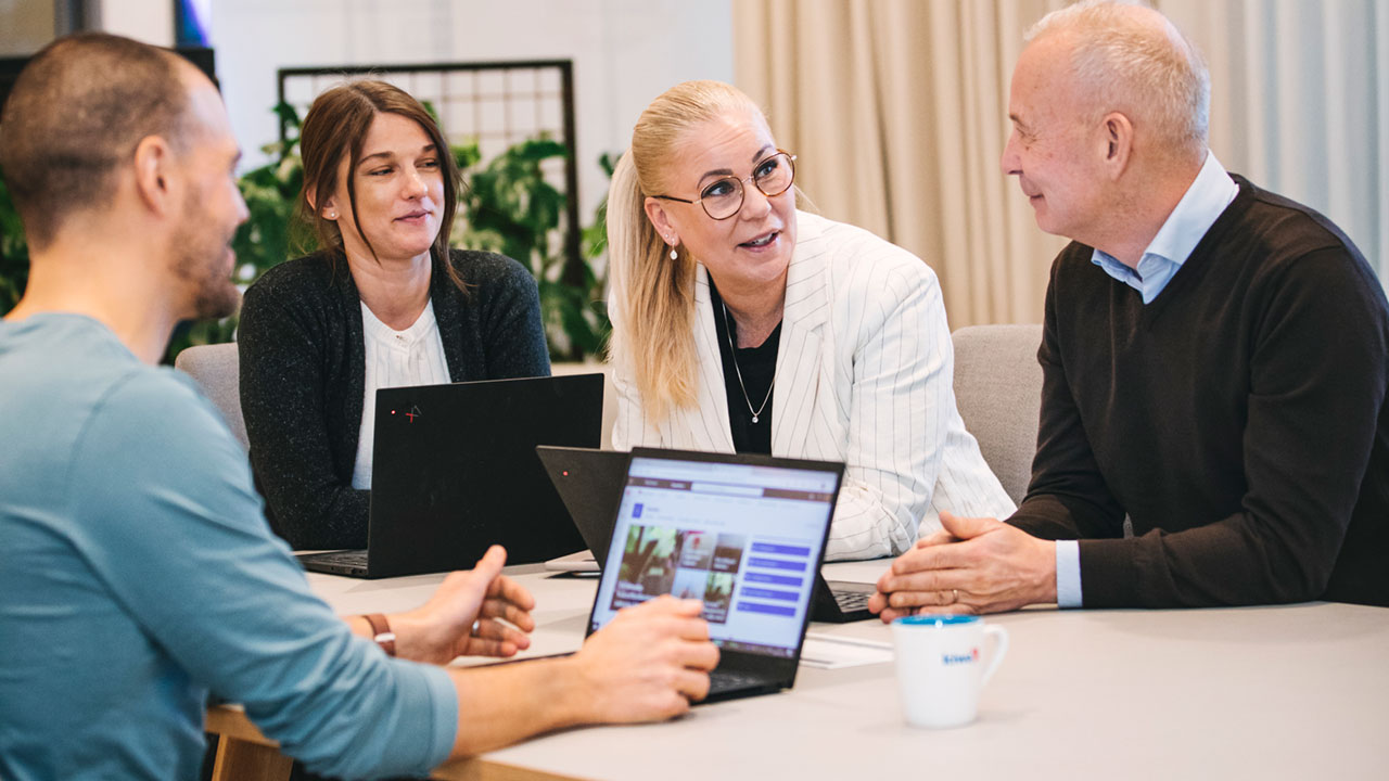 Fyra personer sitter och samtalar vid ett bord i ett kontorsmiljö. Två har laptops framför sig och en vit kaffekopp står på bordet.