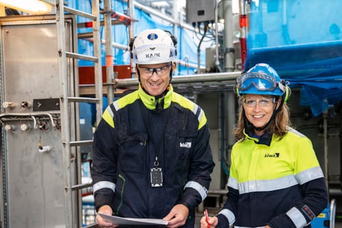 Två besiktningsingenjörer från Kiwa i arbetskläder och skyddshjälmar står på en industriell arbetsplats i bakgrunden.