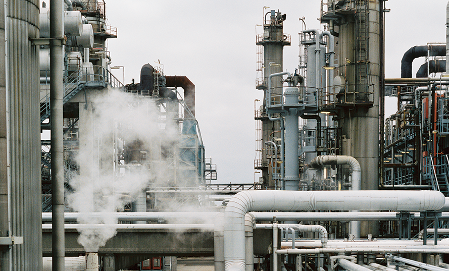 Industriell anläggning med rör, skorstenar och ånga som stiger upp i luften under en molnig himmel.