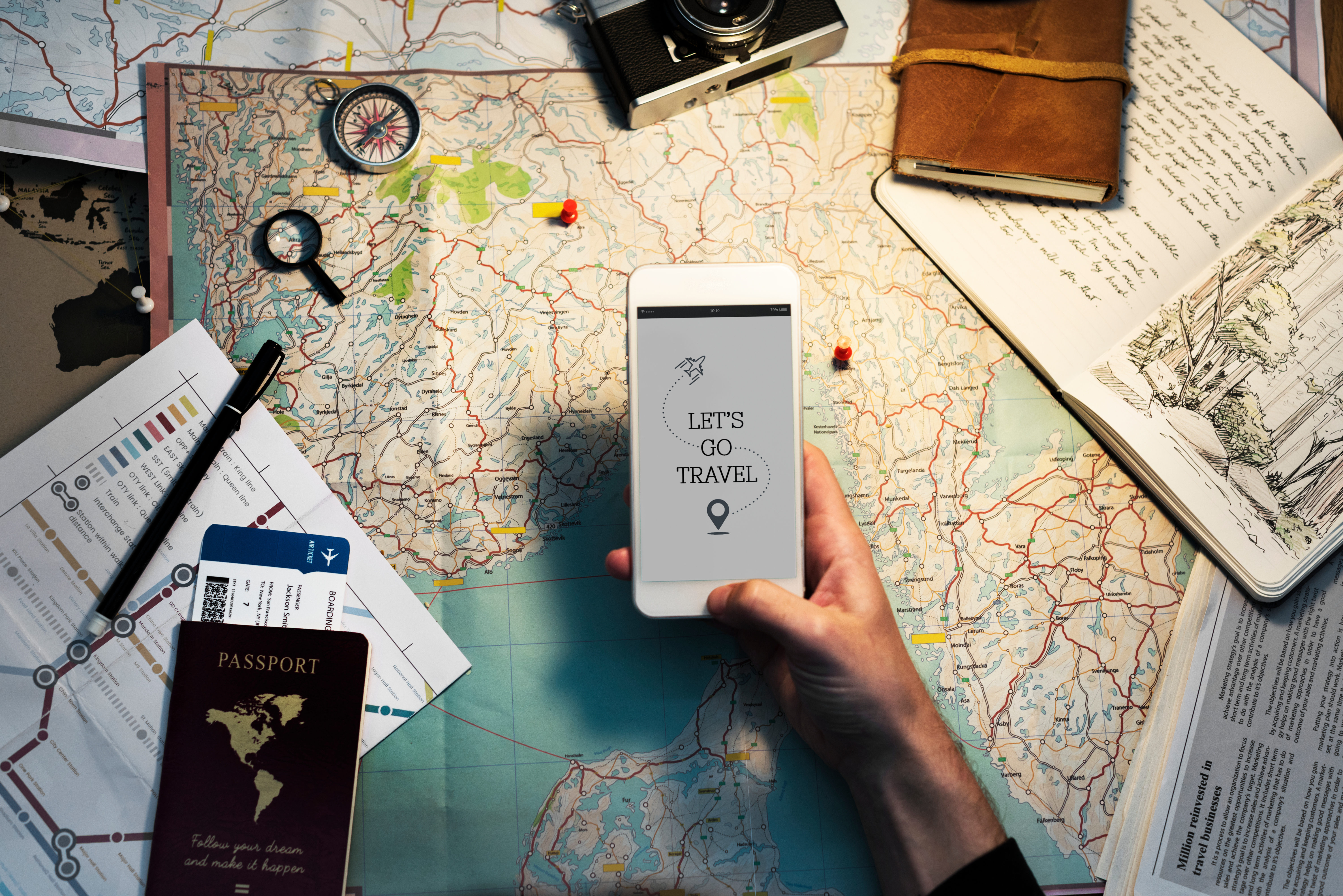Travel planning concept with a person holding a smartphone displaying "Let's Go Travel" over a world map. Surrounding items include a vintage camera, compass, passport, and travel documents, enhancing the theme of adventure and exploration