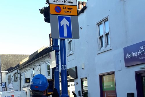 Kiwa employee inspecting street sign in urban area with one-way traffic and parking restrictions, featuring nearby buildings and service van