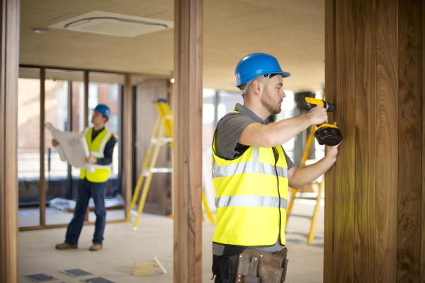 Construction workers in safety vests and helmets using power tools in a modern building interior, with ladders and blueprints in the background