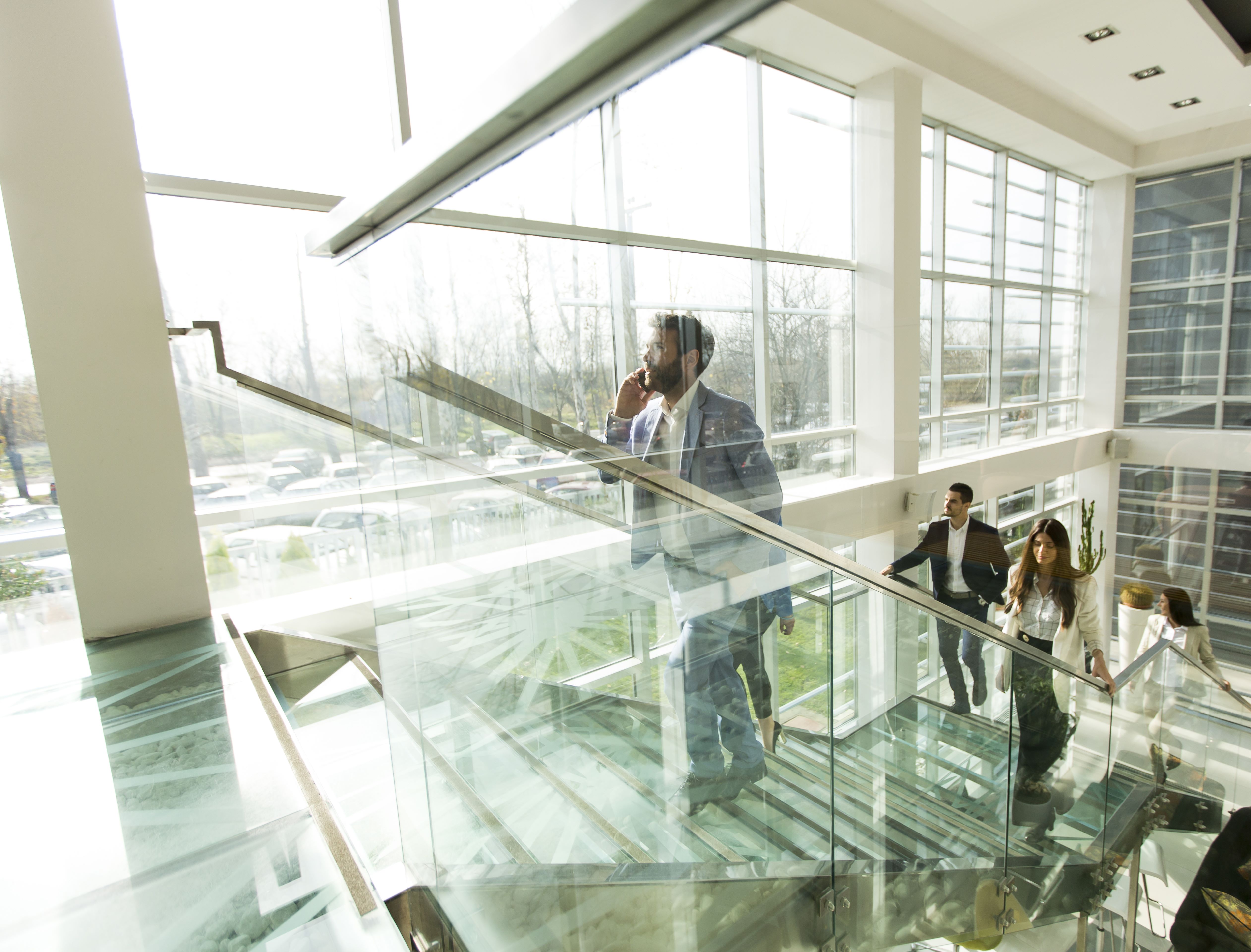 Modern office interior with business professionals walking up a glass staircase, featuring large windows and natural lighting