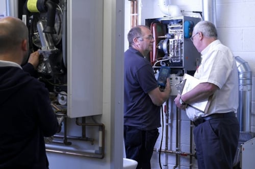 Kiwa trainer and student are inspecting a heating system in a utility room, examining complex piping and machinery