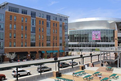 Downtown view of Lincoln, Nebraska featuring Pinnacle Bank Arena, street cafes, and parked vehicles in sunny weather