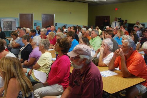 Community members gather in a meeting room for a local public discussion, seated at tables and attentively listening to speakers